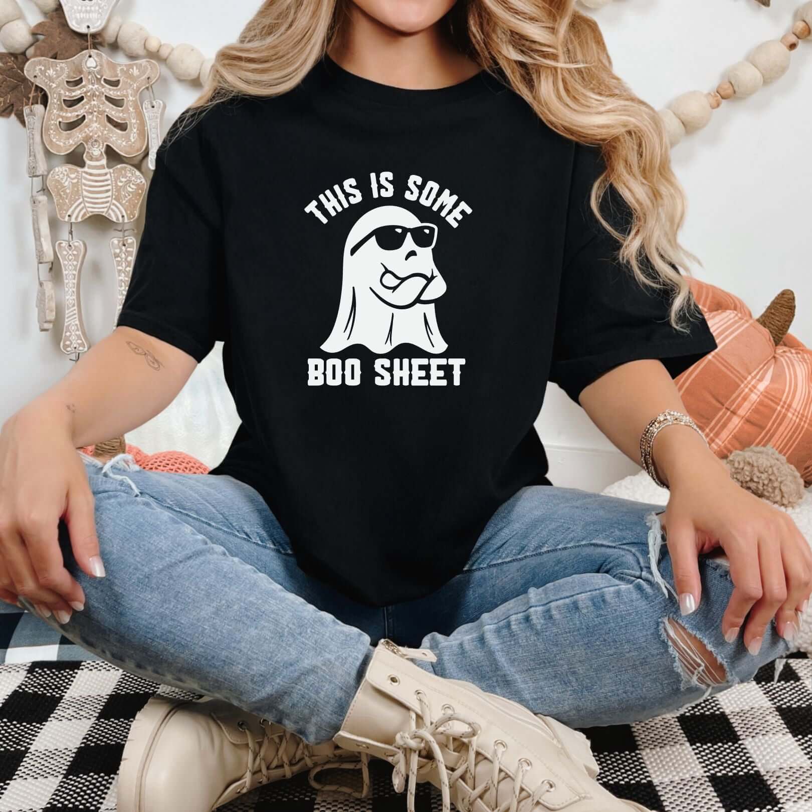 Woman wearing black "This is Some Boo Sheet" graphic tee, sitting cross-legged with pumpkin decorations in the background