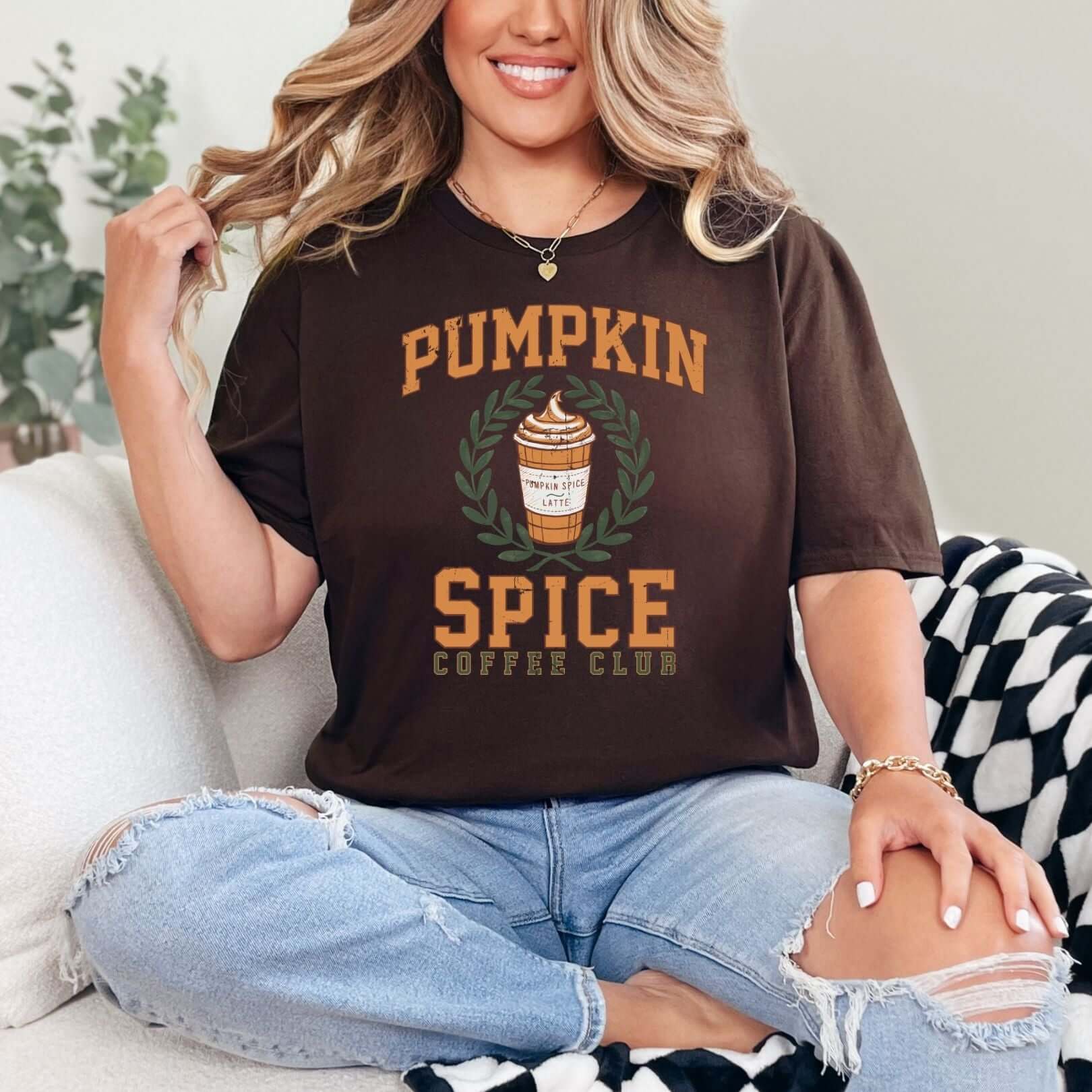 Woman wearing dark brown Take Me to the Pumpkin Patch Graphic Tee with Pumpkin Spice Latte design, sitting on a couch in ripped jeans