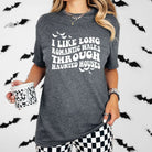 Woman wearing dark grey "Long Haunted Walks" graphic tee with bats in background, holding a patterned mug. Unisex fit, multicolor print.