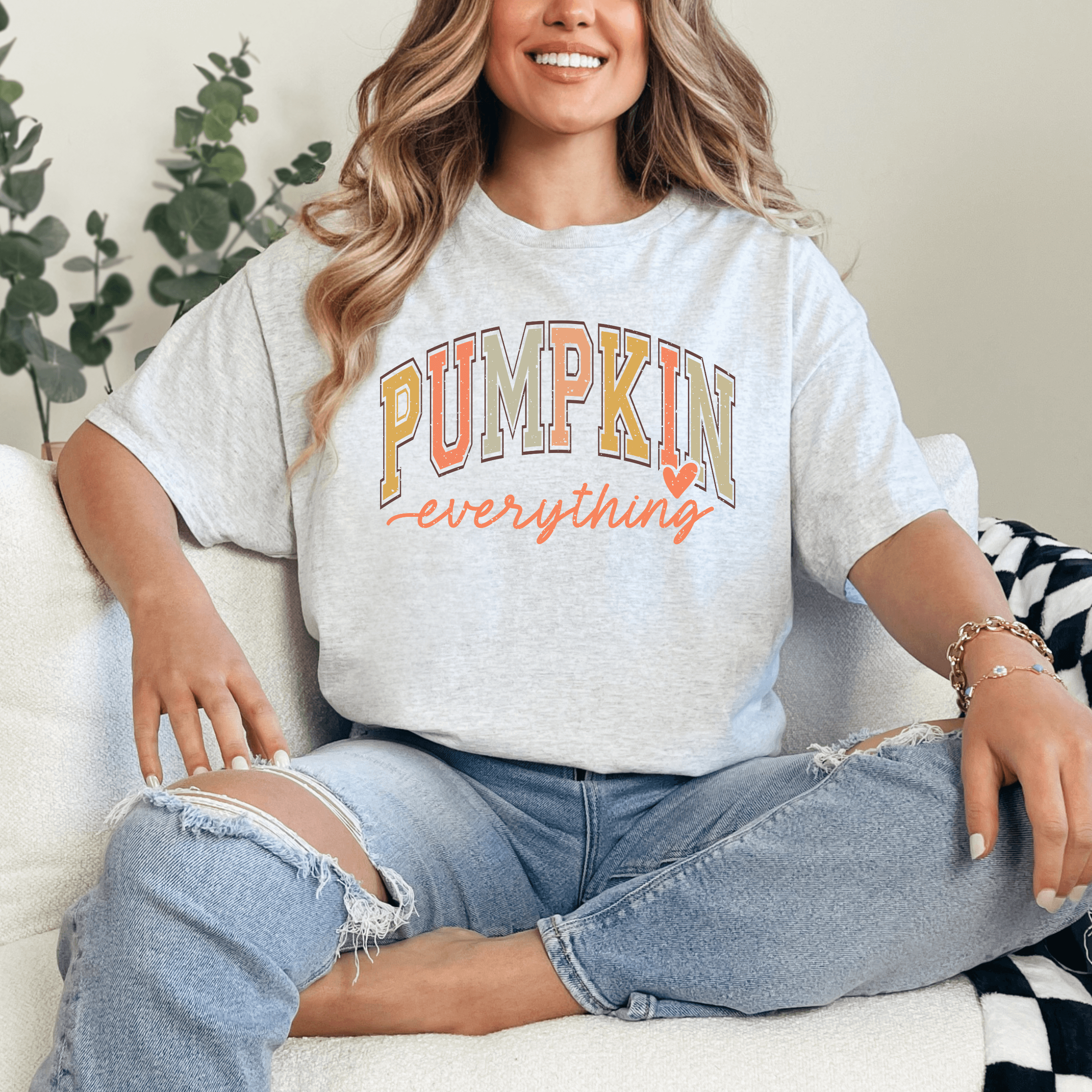 Woman wearing light heathered grey Pumpkin Everything graphic tee with multicolor DTF printing, sitting on a white couch.