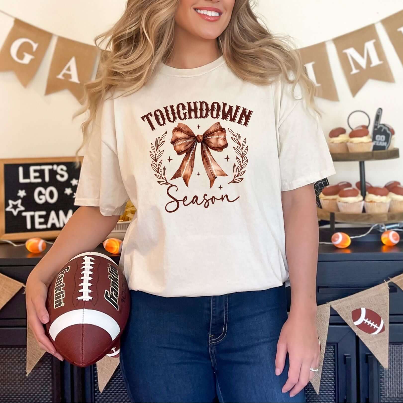 Woman wearing Touchdown Season graphic tee holding a football in front of a football-themed party setup.