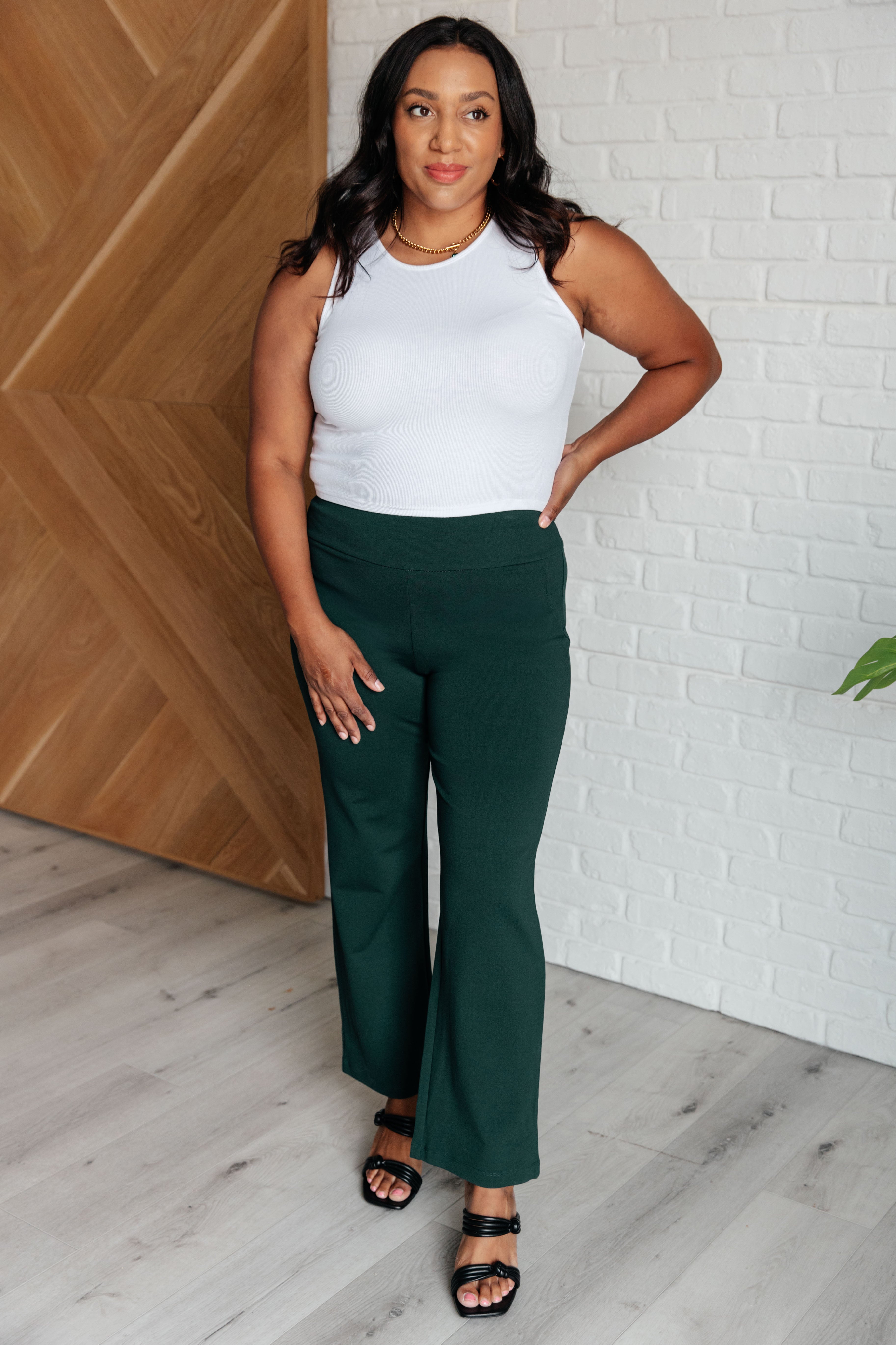 Woman wearing Magic Straight Pants in Hunter Green with white top standing against wooden and brick wall background.