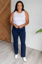 Woman wearing Magic Straight Pants in Navy with a white tank top and white boots, standing in a room with wooden and brick accents.